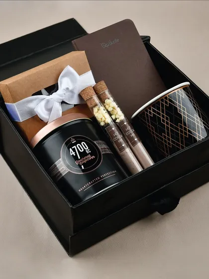 The 'Cozy Vibes' gift hamper is presented in a luxurious black magnetic box. It contains gourmet popcorn, hot chocolate tubes, a notebook, and a stylish mug.