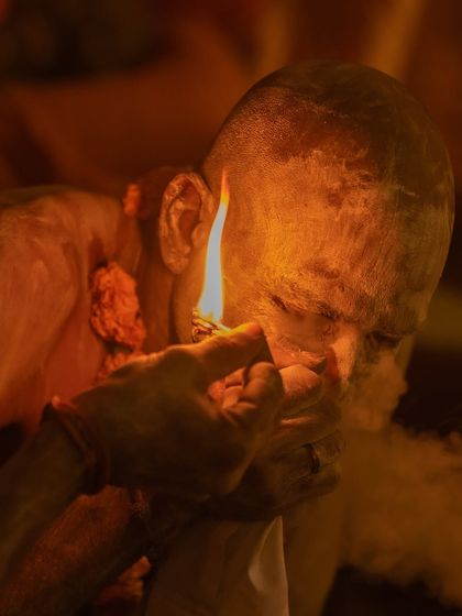 A close-up, dramatic shot of a sadhu lighting his chillum at the Kumbh Mela. The flame illuminates his face, creating an intense and spiritual portrait.