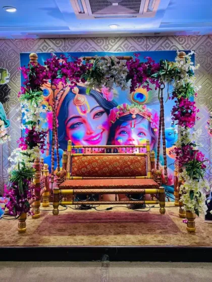 A beautiful and divine setup for a baby's naming ceremony or 'dohale jevan'. This stage features a decorated floral jhula (cradle) with a backdrop of Lord Krishna and Yashoda, surrounded by character cutouts and floral arrangements.
