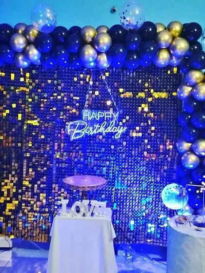 A black and gold sequin wall with a blue and gold balloon arch, lit by a blue spotlight.