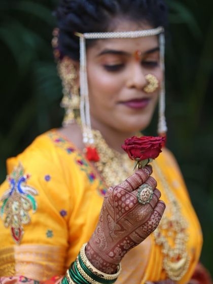 A close-up of the bride's hand, holding a single red rose. The dark henna stain provides a beautiful backdrop for her wedding jewelry.