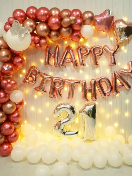 A beautiful 21st birthday decoration in rose gold and white. The setup features a balloon arch against a wall of fairy lights, with large '21' foil balloons.
