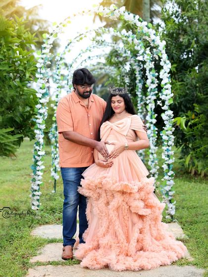 A regal portrait of a mother-to-be in a peach ruffled gown and crown, standing under a floral archway.
