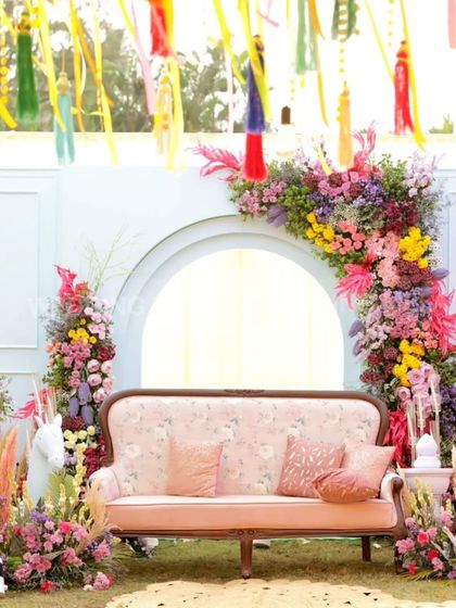 A full view of a whimsical mehandi stage. The pastel pink sofa, vibrant floral arch, and colorful tassels hanging from the ceiling create a fun and dreamy setting.