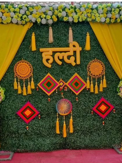 A Haldi backdrop with an artificial green wall, decorated with a "Haldi" sign, dreamcatchers, and colorful hangings. The yellow drapes on the sides complete the look.