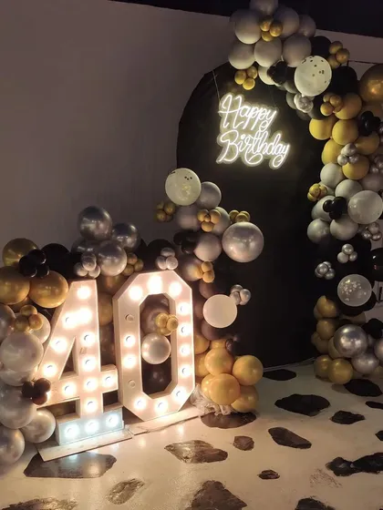 A bold and stylish 40th birthday decoration. The black, silver, and gold balloon arch is paired with a dark backdrop, a neon sign, and large marquee numbers.