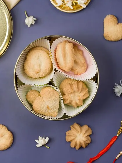 A top-down flat lay of assorted cookies in a tin box, surrounded by Rakshabandhan elements. This composition is visually balanced and tells a festive story.