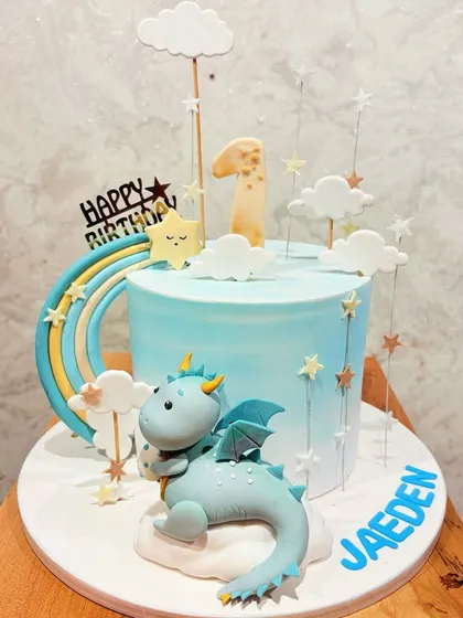 A magical cake for a first birthday, featuring a friendly blue dragon, a rainbow, and dreamy clouds.
