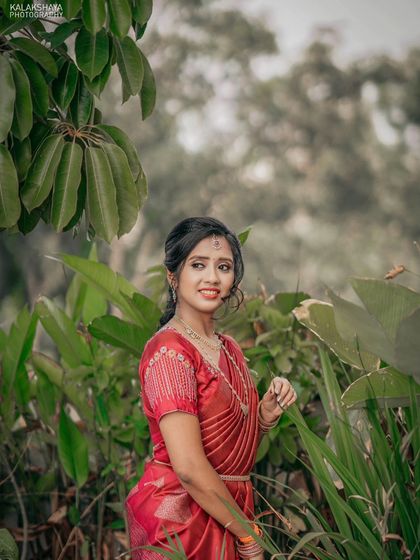 A candid portrait of a bride in a red saree, surrounded by lush green foliage.