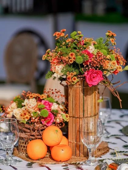 A rustic centerpiece with oranges, chrysanthemums, and roses in a bamboo-wrapped vase, perfect for a summer-themed event.