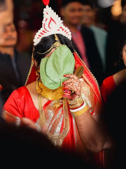 A traditional moment from a Bengali wedding. The bride performs the 'shubho drishti' ritual, hiding her face behind betel leaves before the first look, creating a sense of beautiful anticipation.