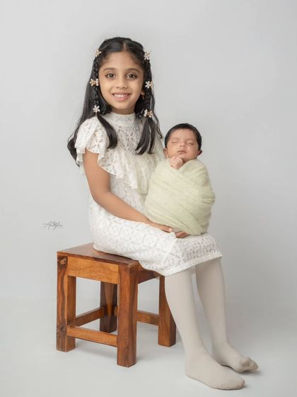 A proud older sister sits on a stool, holding her swaddled newborn sibling on her lap in a clean, simple studio setting.
