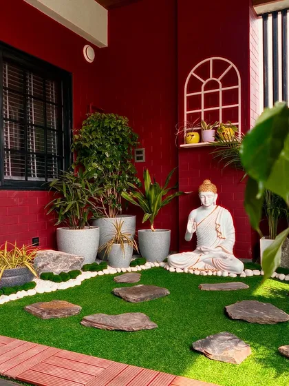 A balcony in Faridabad transformed into a tranquil Zen garden. The space features a serene Buddha statue, artificial grass with stone pavers, and a variety of potted plants against a bold red wall.