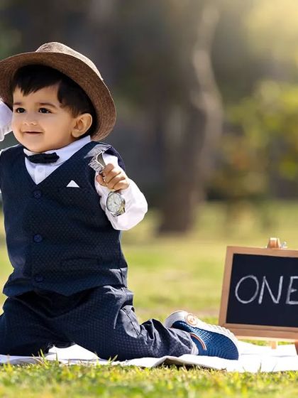A toddler kneels on a blanket in a park, holding a prop watch for his "dapper" themed first birthday shoot.