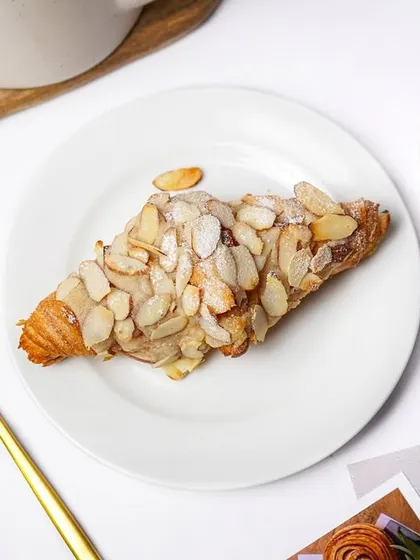 A flaky almond croissant, generously topped with sliced almonds, presented on a white plate with golden cutlery. This is an elegant shot for a high-end bakery or cafe.