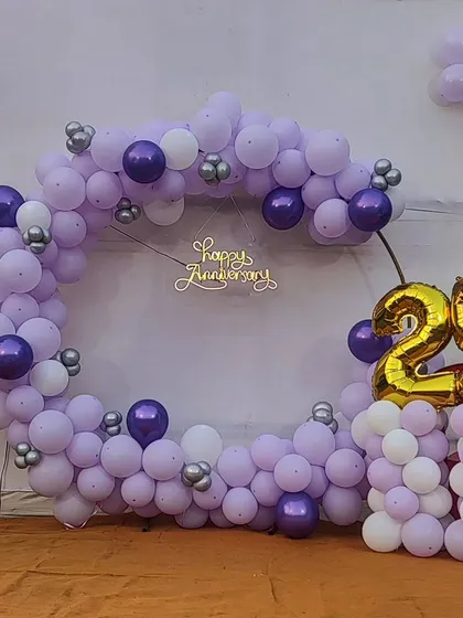 I help celebrate major milestones like this 25th anniversary. This setup includes a circular balloon ring in pastel lavender, white, and chrome purple, a 'Happy Anniversary' neon sign, and a large '25' foil balloon number to mark the special occasion.