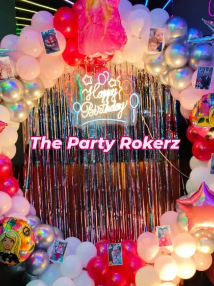 A circular balloon backdrop for a Barbie-themed party. The setup includes a silver fringe curtain, a neon sign, and photos of the birthday girl clipped onto the balloon arch.