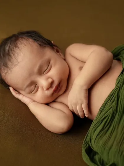 A serene portrait of a newborn sleeping on their side, partially draped with a soft green cloth on an olive green background.