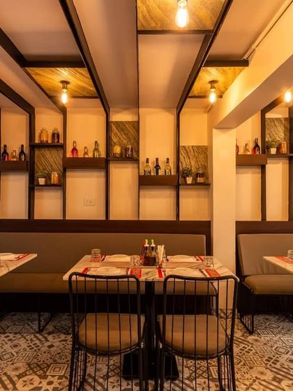A cozy dining nook designed with long booth seating against a wall featuring decorative shelving and vertical wood-finished panels. The exposed Edison bulbs hanging from the ceiling add a warm, vintage touch to this intimate area.