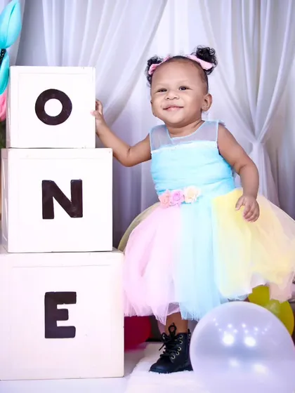 A proud and happy one-year-old girl stands by her birthday blocks in a colorful pastel dress.