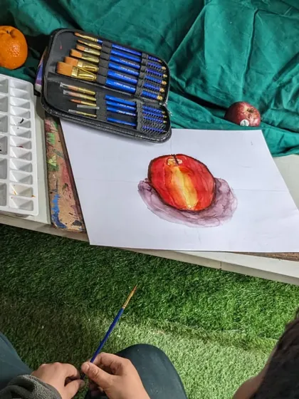 A student's perspective during a still life class. The artist is working on a watercolor painting of an apple, with the reference fruit and art supplies close by.