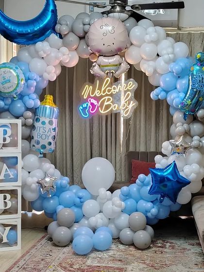 A full circular balloon arch in blue, white, and grey for a baby boy's welcome party. The decor is complete with a neon 'Welcome Baby' sign and 'BABY' letter boxes.