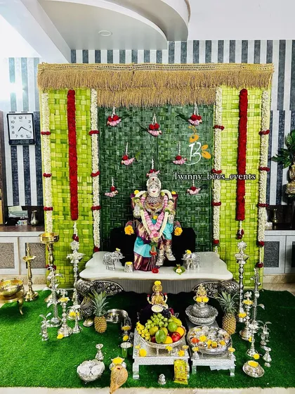 An eco-friendly Varamahalakshmi puja setup featuring a backdrop hand-woven from green coconut palm leaves. The design is accented with red flower hangings and traditional silver lamps, creating a beautiful and sustainable sacred space.