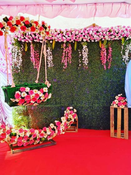 An outdoor naming ceremony setup with a floral crescent moon cradle. The backdrop features a green wall with hanging pink flowers, creating a fresh, garden-like atmosphere.