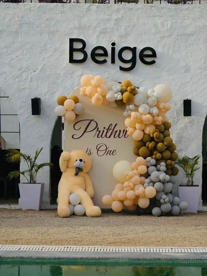 A chic, beige-themed first birthday party at a poolside venue. The decoration features an organic balloon arch in neutral tones, a personalized backdrop, and a large teddy bear prop.