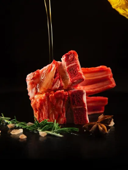 An action shot of olive oil being drizzled over a stack of raw ribs against a black background. The dramatic lighting highlights the texture of the meat and the stream of oil, creating a luxurious and dynamic feel.
