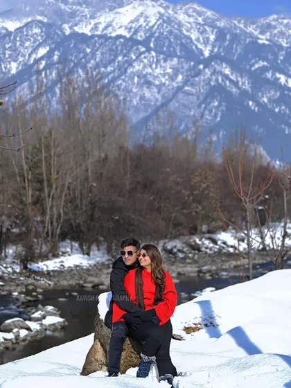 A cozy couple portrait in the snow, with a river and mountains in the background. The vibrant red of her outfit stands out against the winter whites.