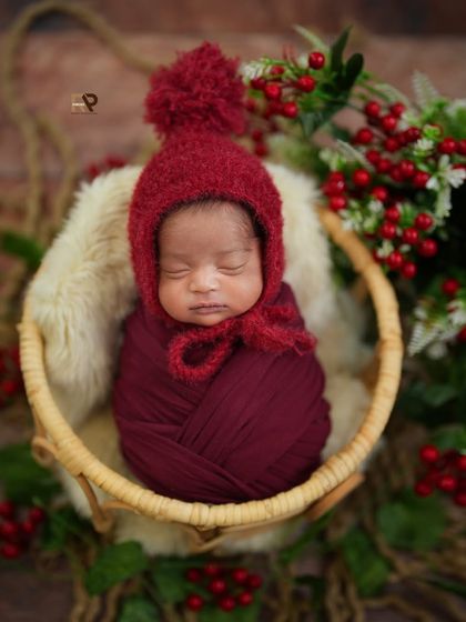 A tiny newborn, wrapped in a cozy red swaddle and wearing a matching bonnet, sleeps peacefully in a rustic wicker basket. The surrounding red berries add a festive, winter-themed touch to the photograph.