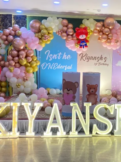 A grand stage setup for a teddy bear themed first birthday party, complete with large marquee letters spelling out the name 'KIYANSH' and multiple themed backdrops.
