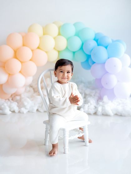 A serene first birthday portrait with a pastel rainbow balloon arch.