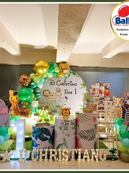 A jungle-themed first birthday setup with a circular backdrop and various animal cutouts. The green and gold balloon arch adds a touch of safari elegance.