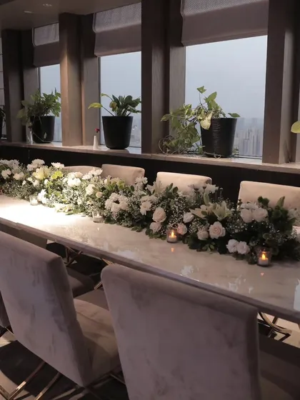An elegant long-table dining setup, featuring a runner of white flowers and greenery, complemented by the warm glow of candles, perfect for an intimate dinner.