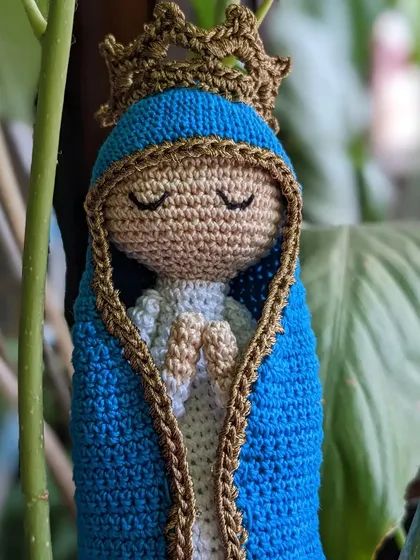 This serene Mother Mary doll is shown nestled amongst leaves, highlighting her peaceful presence. She wears a blue mantle and a golden crown.