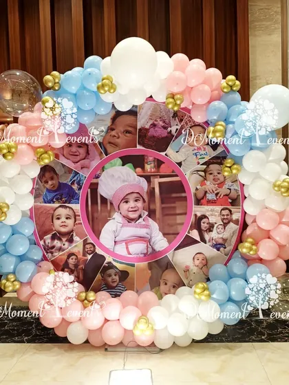 A creative photo collage display for a first birthday. The photos are arranged in a circle and framed by a beautiful balloon garland in pastel pink, blue, and white.