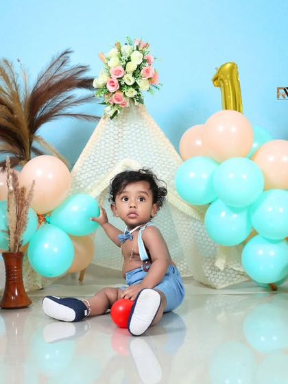 A baby boy sits amidst a charming first birthday setup, featuring a teepee, "ONE" blocks, and a teddy bear. The pastel blue and peach balloons add a soft and dreamy feel.