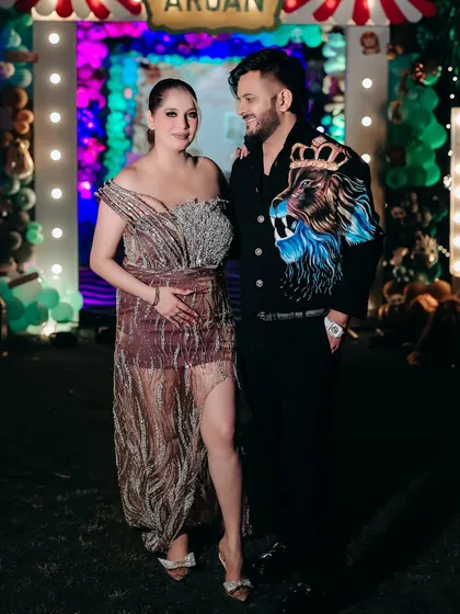A happy couple posing at what appears to be a baby shower or celebration. The mom-to-be cradles her bump in a glamorous dress, surrounded by festive decorations.