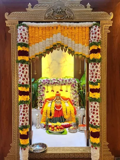 A beautifully decorated pooja room for a housewarming ceremony. The deity is adorned with flower garlands, and the setup includes fruits and other offerings.