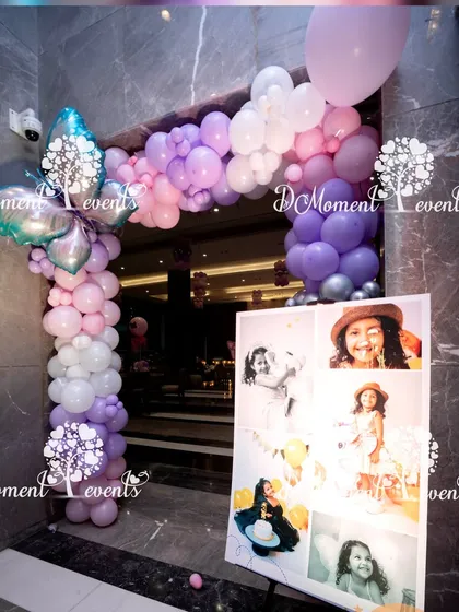An entrance decorated with a pastel purple and pink balloon arch. A photo collage of the birthday girl stands beside it, offering a personal welcome to guests.