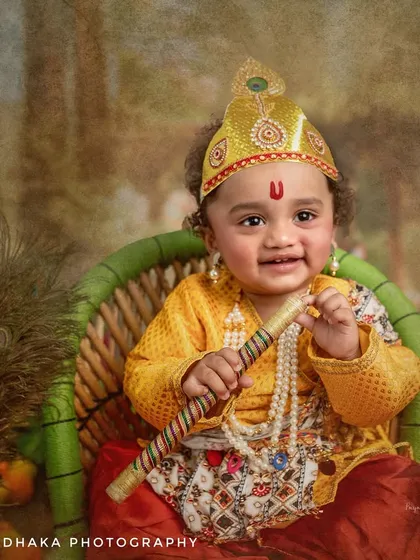 A happy one-year-old Krishna holds his flute, ready to charm the world. The setup includes a wicker chair and peacock feathers for a touch of rustic divinity.