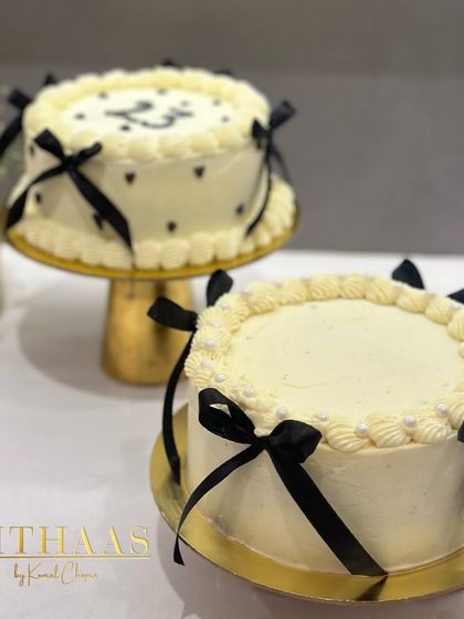 Two beautiful white birthday cakes with black ribbon details. One features small hearts, while the other is simpler, both sitting elegantly on gold cake stands.
