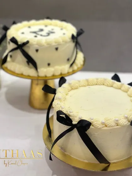 Two beautiful white birthday cakes with black ribbon details. One features small hearts, while the other is simpler, both sitting elegantly on gold cake stands.