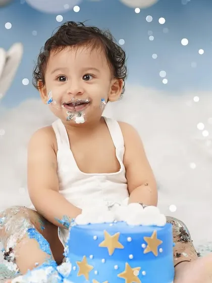 A joyful baby boy with a frosting-covered smile enjoys his first birthday cake smash, set against a dreamy blue backdrop with moon and star props.