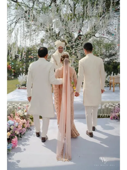 A walk to remember. The bride, escorted by her brothers, makes her way down a pristine white aisle lined with soft pastel flowers, heading towards a beautiful garden mandap for a serene and elegant farmhouse wedding ceremony.