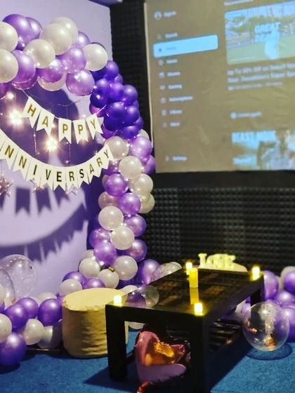 A beautiful anniversary decoration featuring a purple and white balloon arch and a 'Happy Anniversary' banner. The table is set with LED candles for a romantic dinner feel.