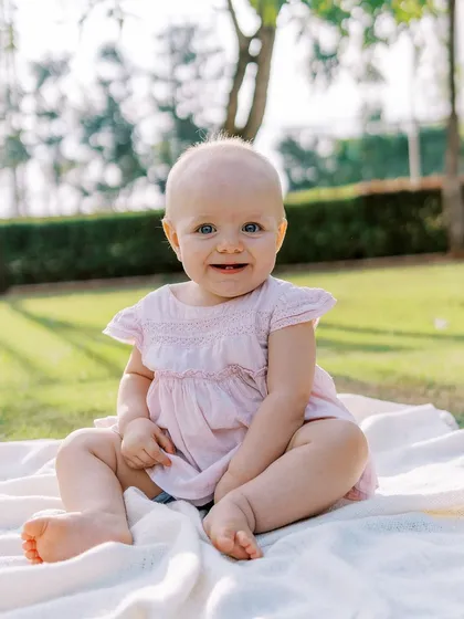 A happy baby girl with a sweet, toothy smile, sitting on a blanket outdoors. Her joy is absolutely radiant.
