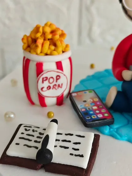 A close-up of the tiny, handcrafted details on the personalized hobby cake, including a miniature popcorn box, an iPhone, and a book with a pen.
